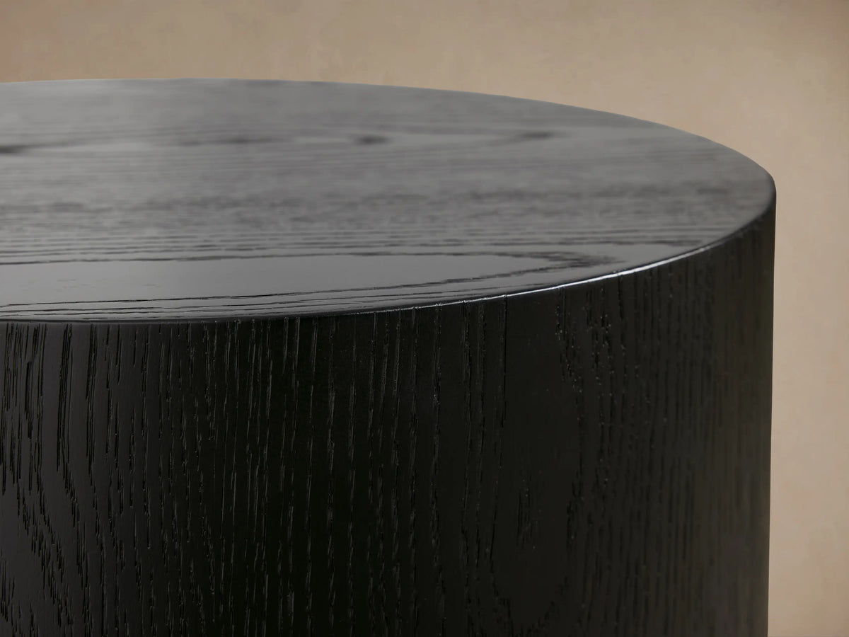 Sullivan Round End Table in black oak finish, cylindrical shape, smooth matte surface, on wooden floor against beige wall.