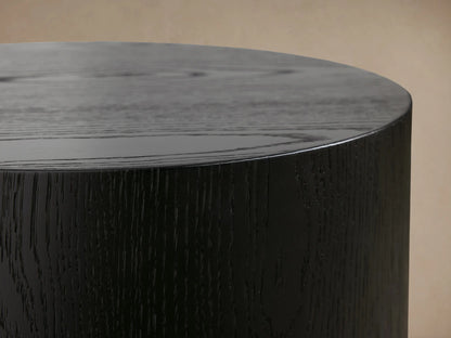 Sullivan Round End Table in black oak finish, cylindrical shape, smooth matte surface, on wooden floor against beige wall.