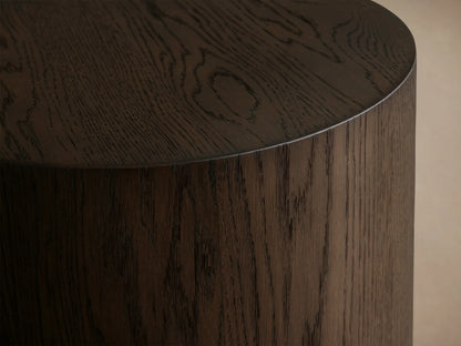 Dark oak cylindrical Sullivan Round End Table with rich wood grain and black plinth base, standing on a light wooden floor.