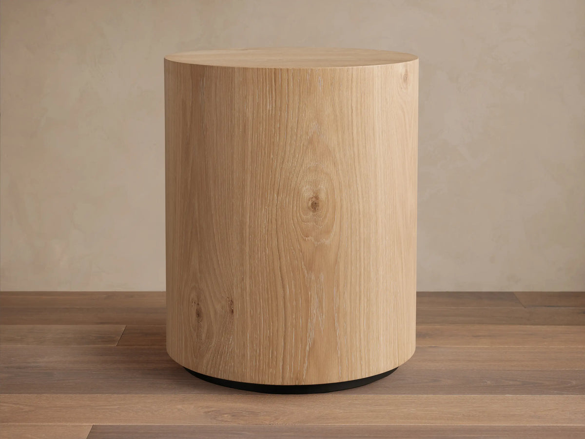 Sullivan Round End Table made of solid oak with a natural grain finish, cylindrical shape, on a black plinth-style base.