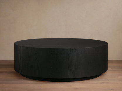 Sullivan Round Coffee Table with a sleek, black, circular design. Oak base with visible wood grain texture on a wooden floor.