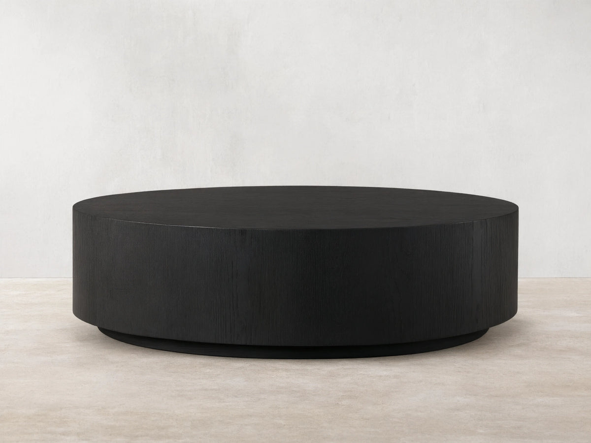 Sullivan Round Coffee Table with a sleek, black, circular design. Oak base with visible wood grain texture on a wooden floor.