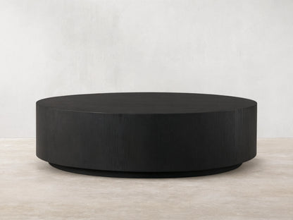 Sullivan Round Coffee Table with a sleek, black, circular design. Oak base with visible wood grain texture on a wooden floor.