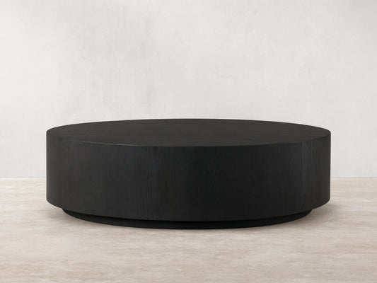 Sullivan Round Coffee Table with a sleek, black, circular design. Oak base with visible wood grain texture on a wooden floor.