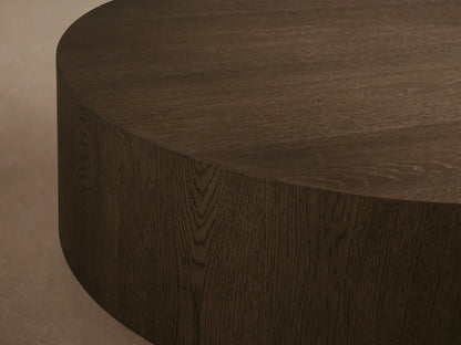 Front view of the light oak Sullivan Round Coffee Table, featuring a black plinth base and decorative items on top.