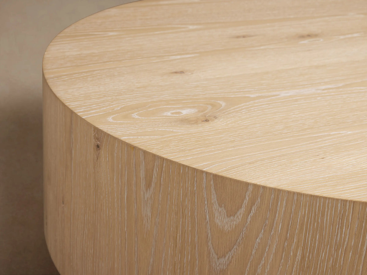Front view of light oak Sullivan Round Coffee Table with a black plinth base, topped with a white vase and open book.