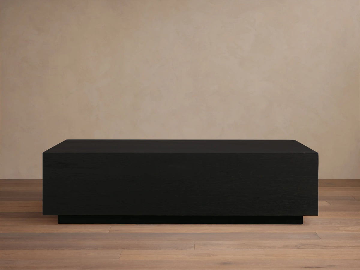 Rectangular Sullivan Coffee Table in dark oak with smooth finish. Sits on a black plinth-style base, against a light beige wall.
