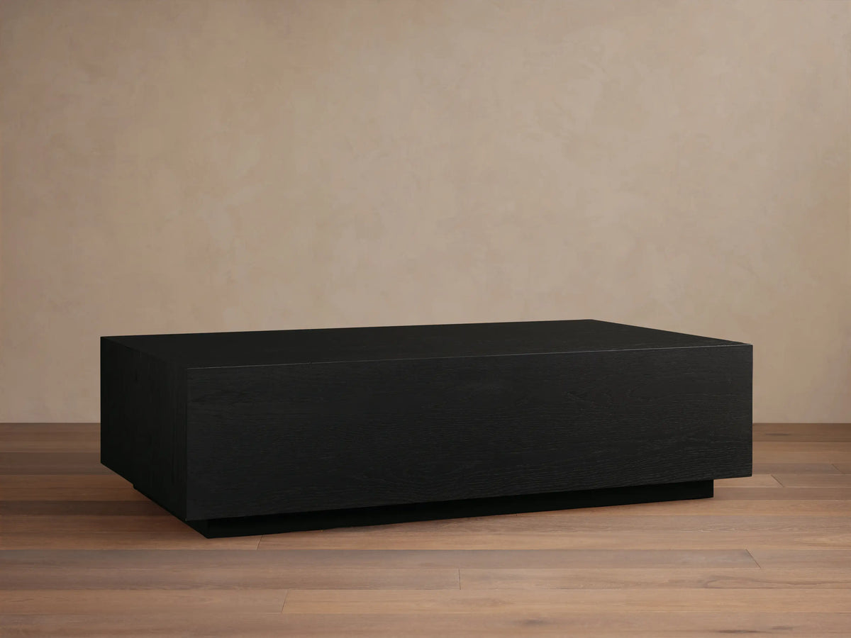 Rectangular Sullivan Coffee Table in dark oak with smooth finish. Sits on a black plinth-style base, against a light beige wall.