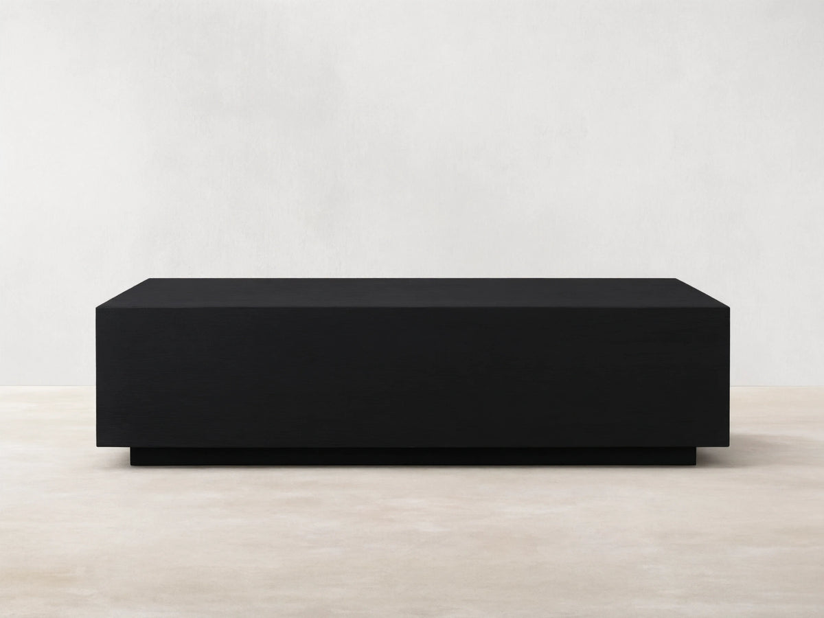 Rectangular Sullivan Coffee Table in dark oak with smooth finish. Sits on a black plinth-style base, against a light beige wall.