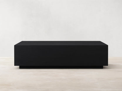 Rectangular Sullivan Coffee Table in dark oak with smooth finish. Sits on a black plinth-style base, against a light beige wall.