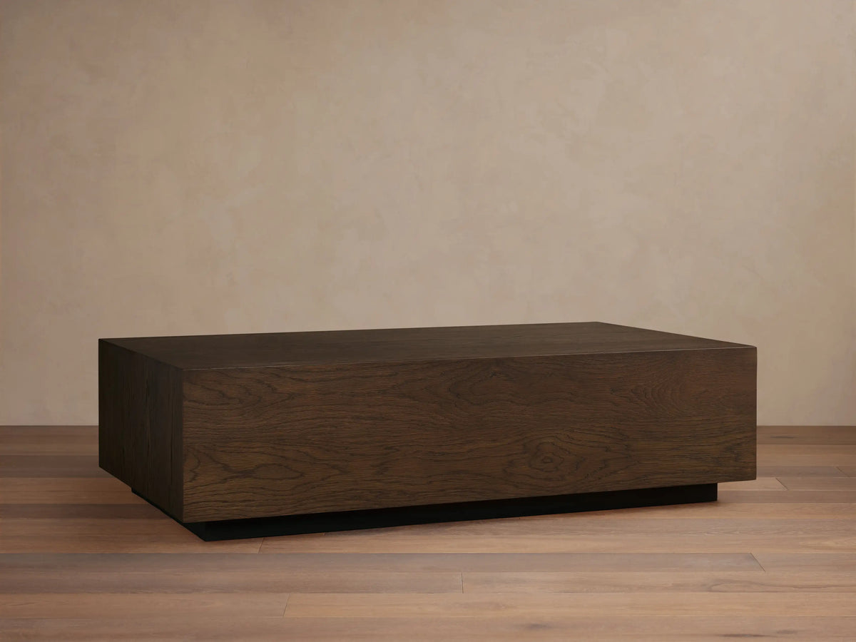 Sullivan Coffee Table in solid oak with visible grain patterns, resting on a black plinth base against a neutral wall.