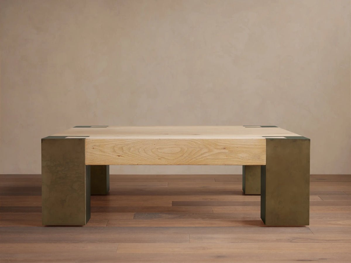 Light oak square Telluride coffee table with four legs of oxidized steel with dark patinas, against a beige wall and wood floor.