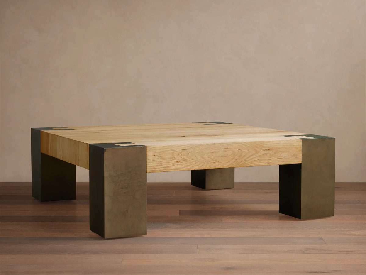 Light oak square Telluride coffee table with four legs of oxidized steel with dark patinas, against a beige wall and wood floor.