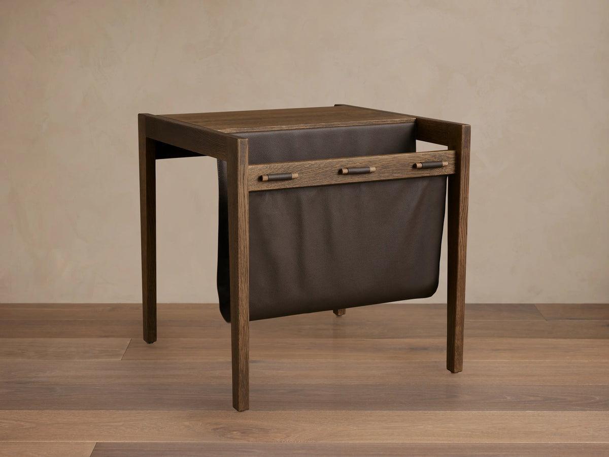 Thorin Magazine Table made of solid oak with a hand-applied finish. Features a dark leather sling on the side for storage.