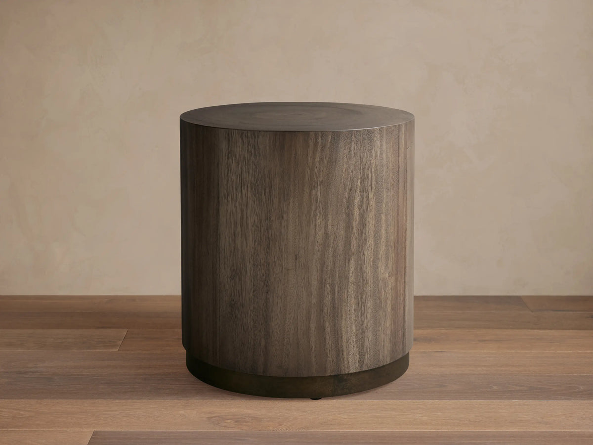 Toluca End Table with a cylindrical shape, dark wood finish, and smooth surface, placed on wooden floor against a light wall.