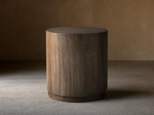 Toluca End Table with a cylindrical shape, dark wood finish, and smooth surface, placed on wooden floor against a light wall.