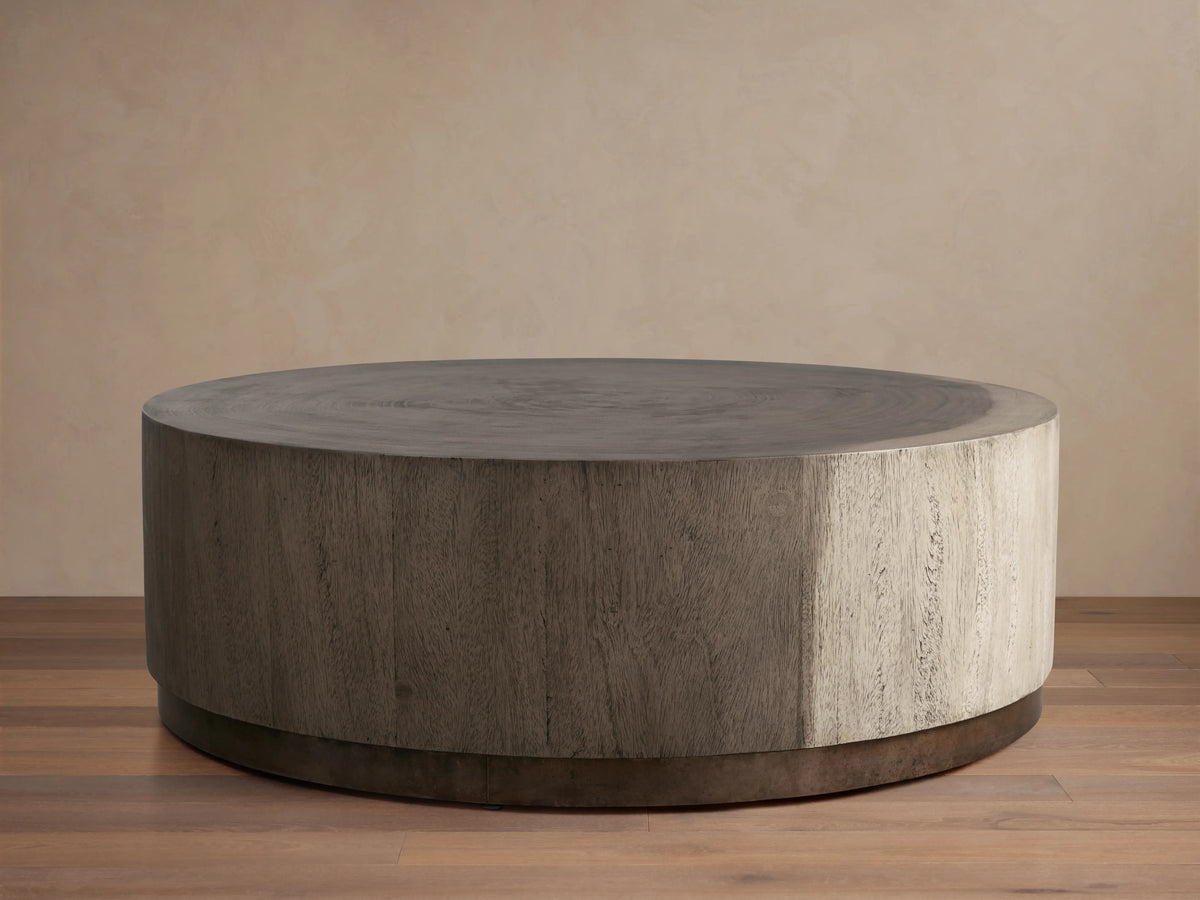Round Toluca Coffee Table, weathered grey guanacaste wood grain top & sides, dark oxidized metal base on wood floor.