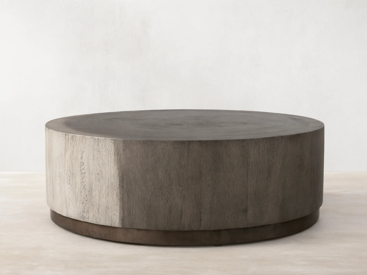 Round Toluca Coffee Table, weathered grey guanacaste wood grain top & sides, dark oxidized metal base on wood floor.