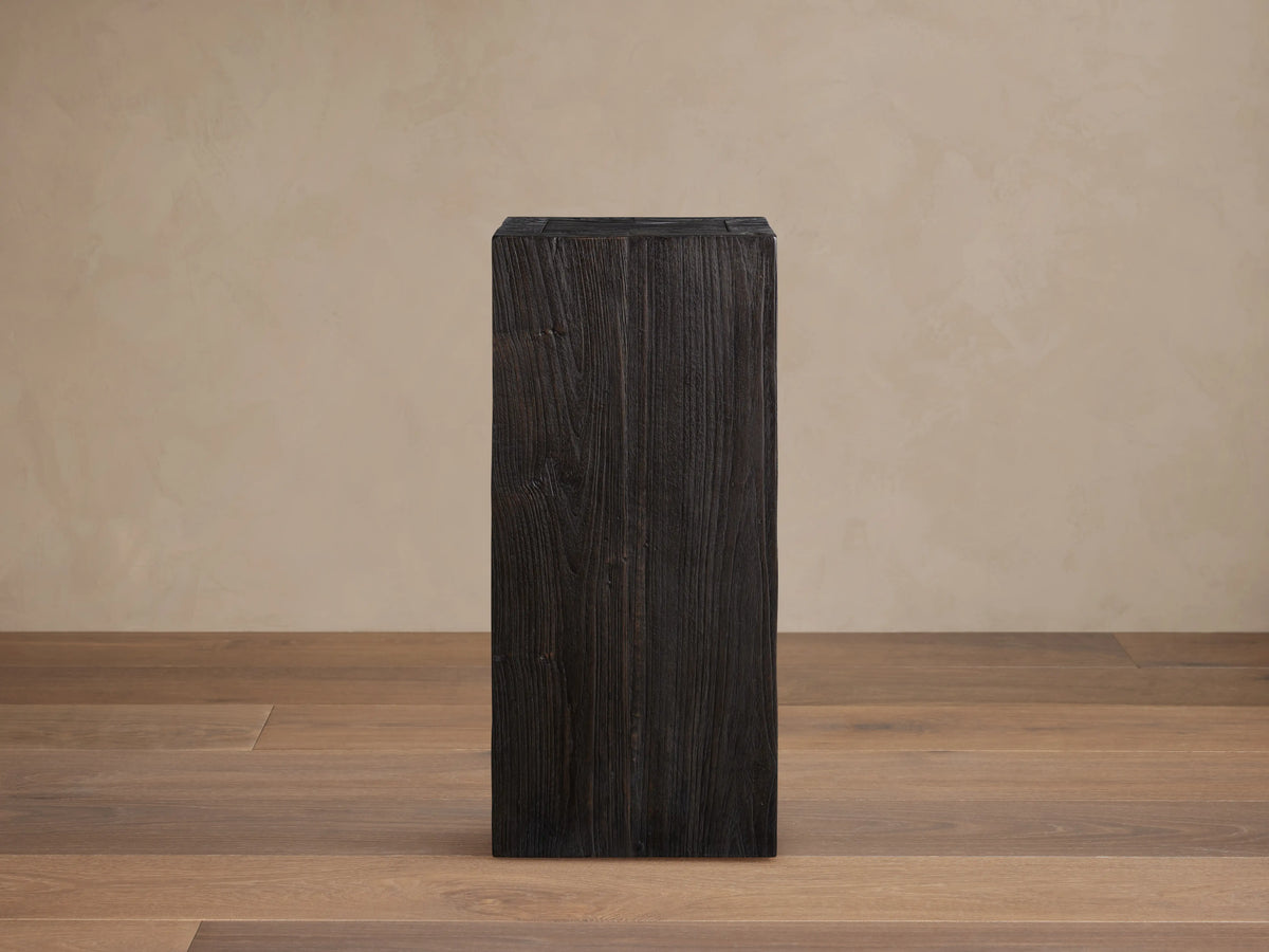 Tall, rectangular Ubud Pedestal made of teak wood with a dark, charred finish and visible grain patterns, set on a wooden floor background.