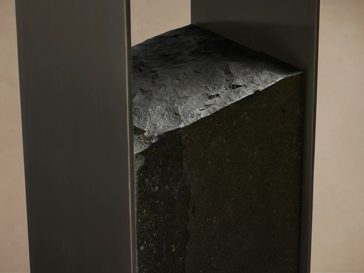 Close-up of Urben Martini Table base: dark, rough-cut marquina marble block with jagged top edge nestled between sleek dark brass supports.