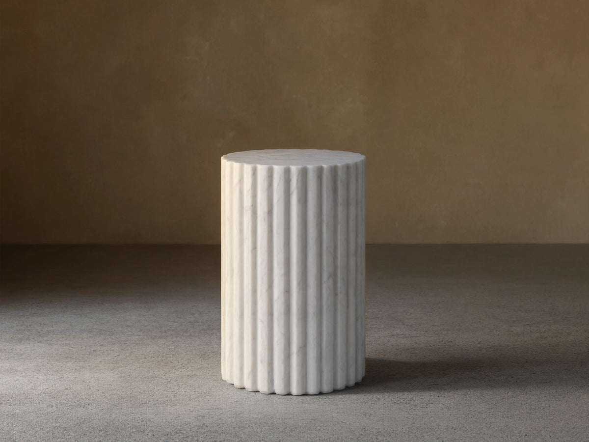 Front view of a white marble fluted cylindrical pedestal on a brown wooden floor.