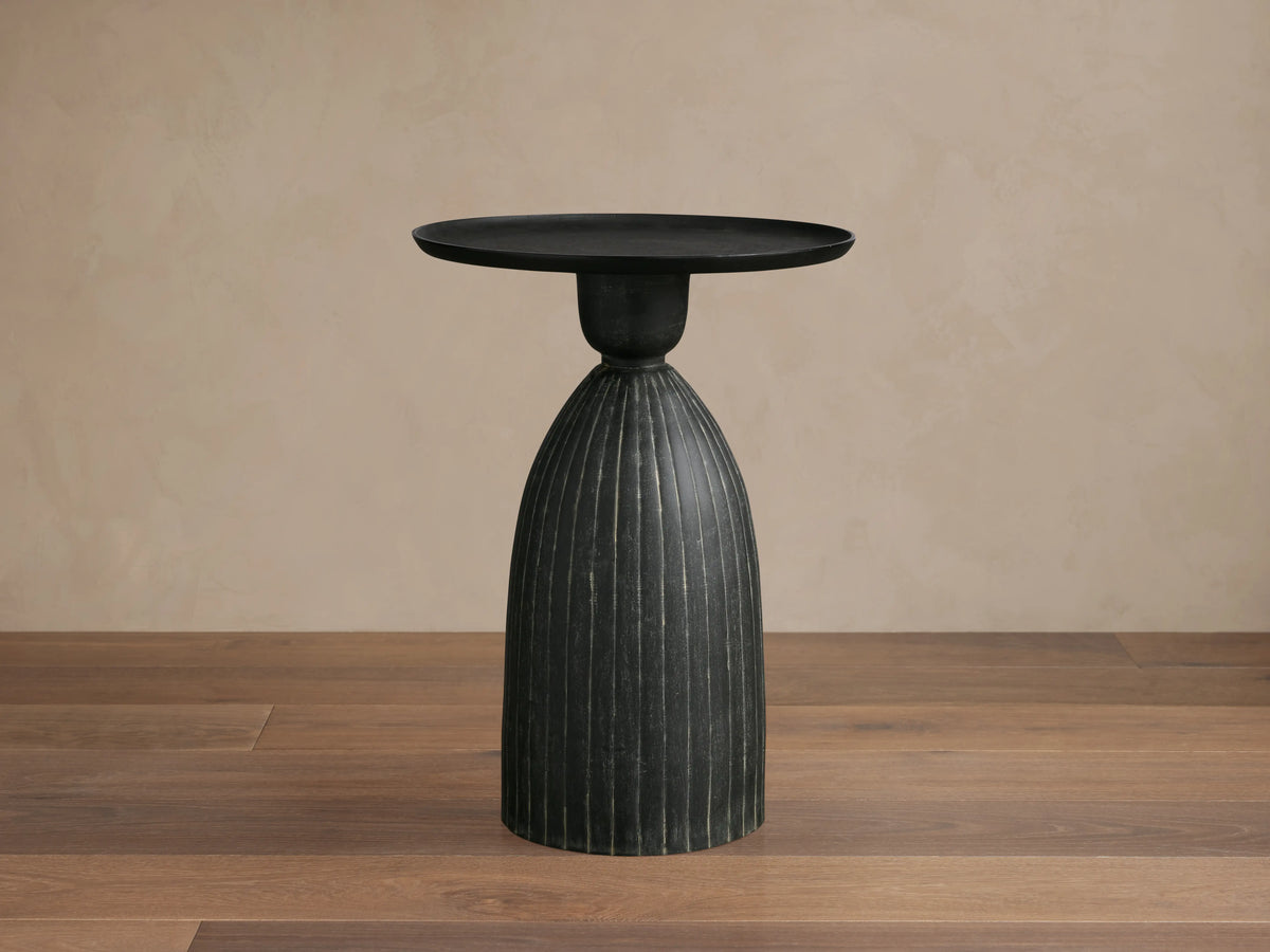 Zephyr Martini Table with black ribbed cast aluminum base and smooth round top on wooden floor, set against a beige background.