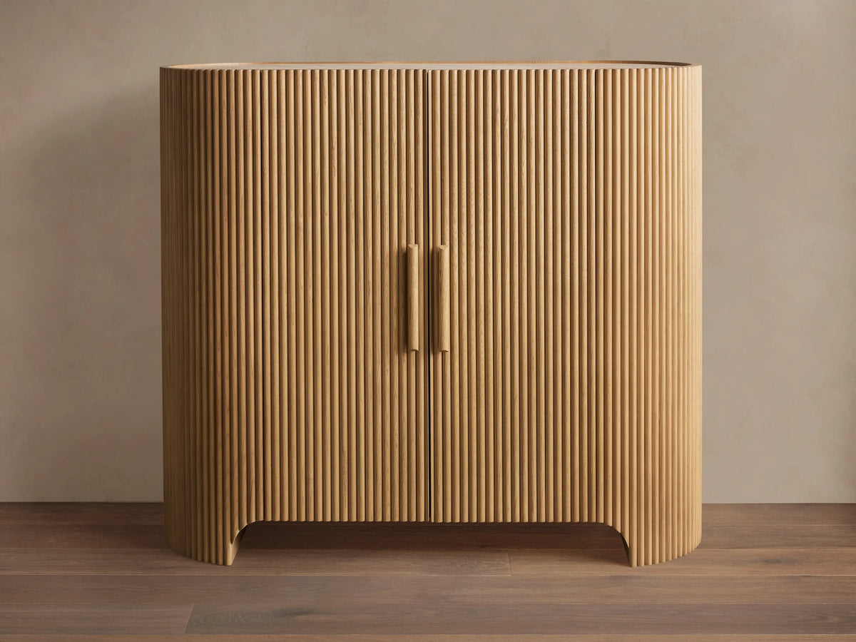 Astor Hall Chest, light oak wood cabinet with vertical reeding, rounded sides, two doors, and wooden pull handles, on wooden floor.