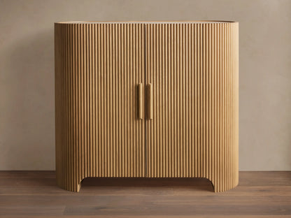 Astor Hall Chest, light oak wood cabinet with vertical reeding, rounded sides, two doors, and wooden pull handles, on wooden floor.