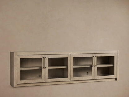 Bodhi Media Console in solid white oak with four glass-front doors, brass handles, and adjustable shelves on a beige backdrop.