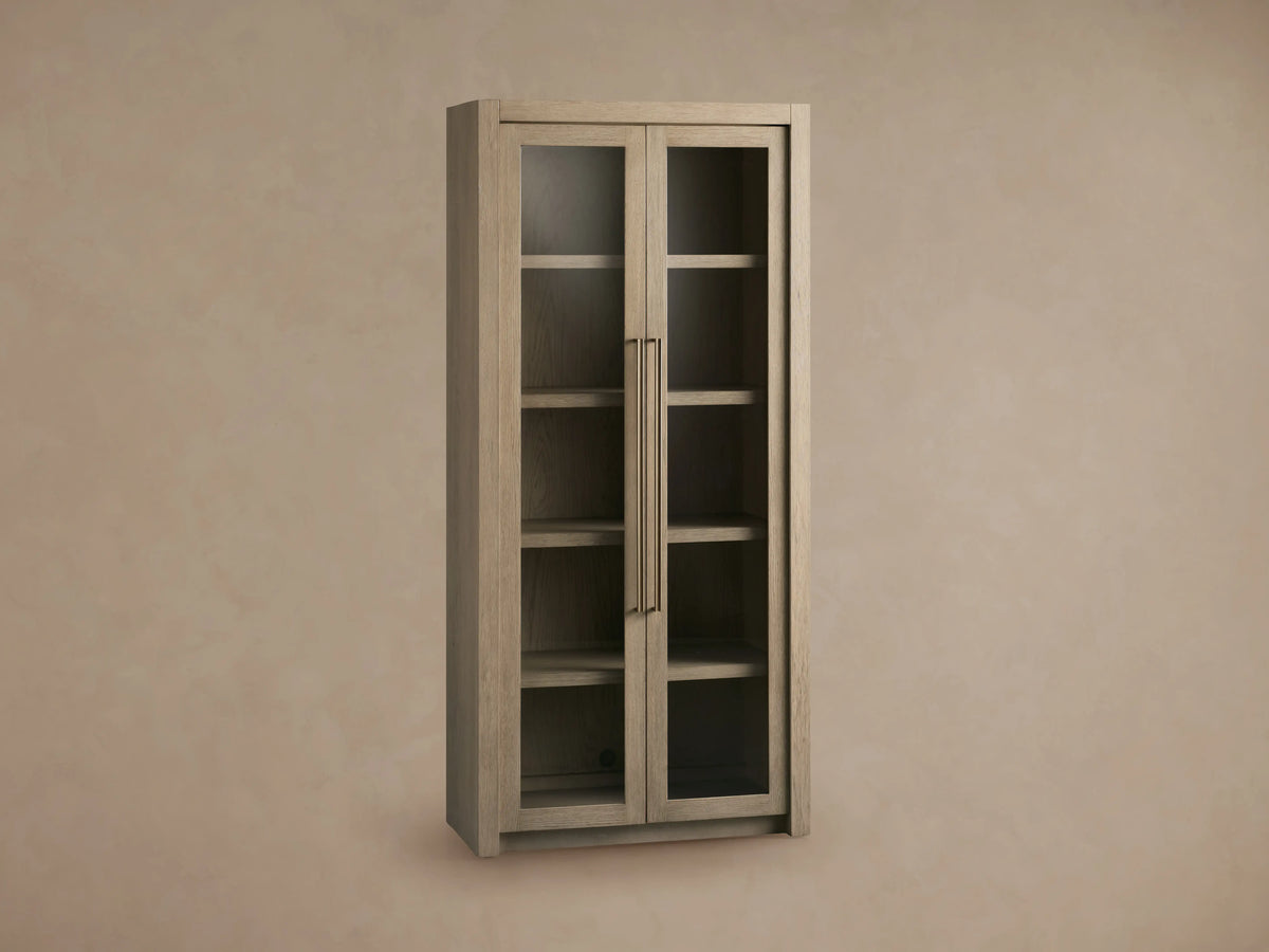 Bodhi Cabinet made of white oak with two glass-panel doors, vertical brass handles, and four visible shelves, on beige background.