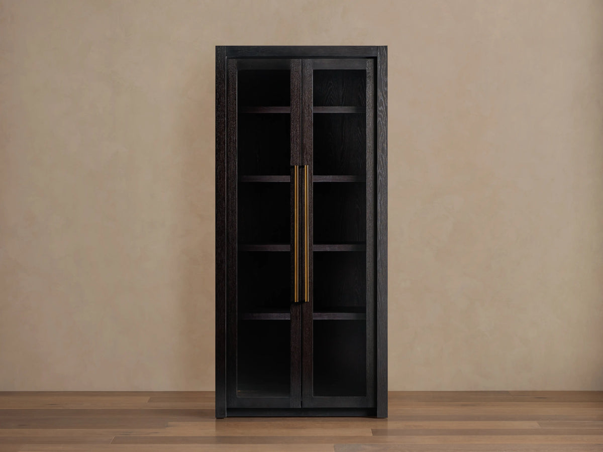 Bodhi Cabinet in dark wood with two glass-paneled doors, gold vertical handles, and a tall, rectangular shape. Placed against a beige wall.