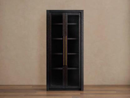 Bodhi Cabinet in dark wood with two glass-paneled doors, gold vertical handles, and a tall, rectangular shape. Placed against a beige wall.