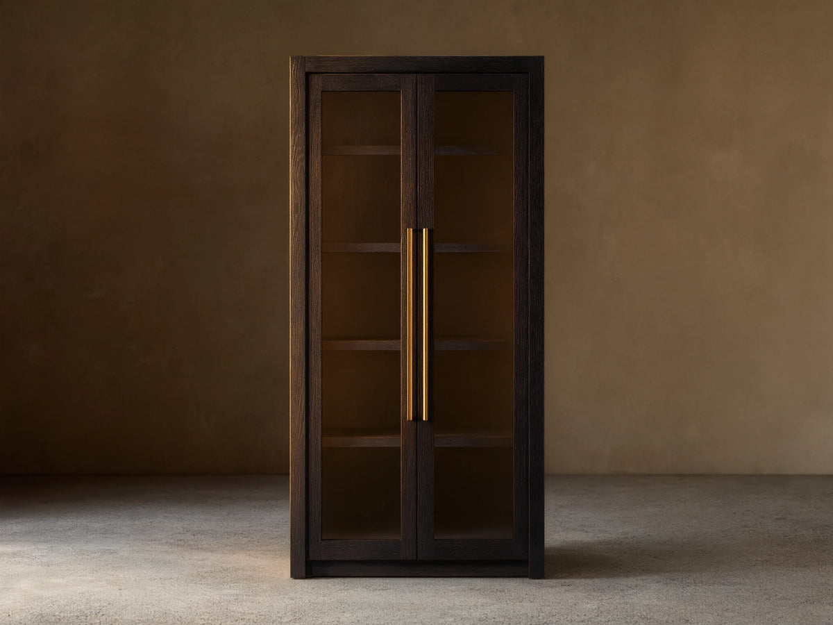 Bodhi Cabinet in dark wood with two glass-paneled doors, gold vertical handles, and a tall, rectangular shape. Placed against a beige wall.
