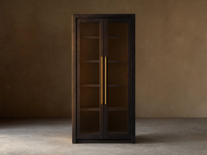 Bodhi Cabinet in dark wood with two glass-paneled doors, gold vertical handles, and a tall, rectangular shape. Placed against a beige wall.