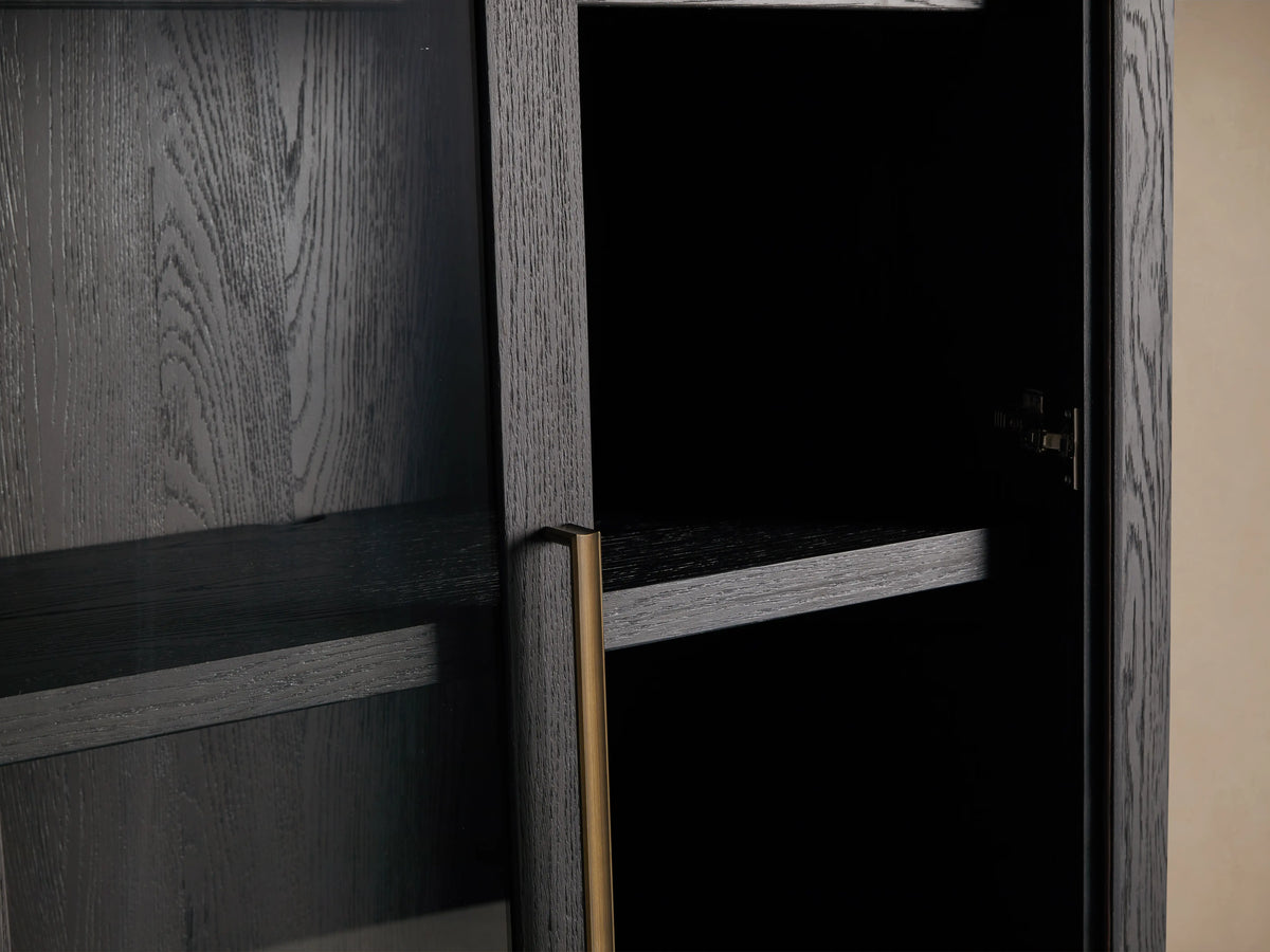 Bodhi Cabinet in dark wood with two glass-paneled doors, gold vertical handles, and a tall, rectangular shape. Placed against a beige wall.