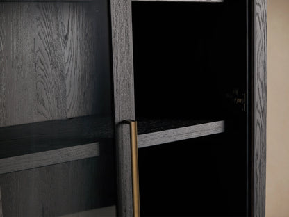 Bodhi Cabinet in dark wood with two glass-paneled doors, gold vertical handles, and a tall, rectangular shape. Placed against a beige wall.
