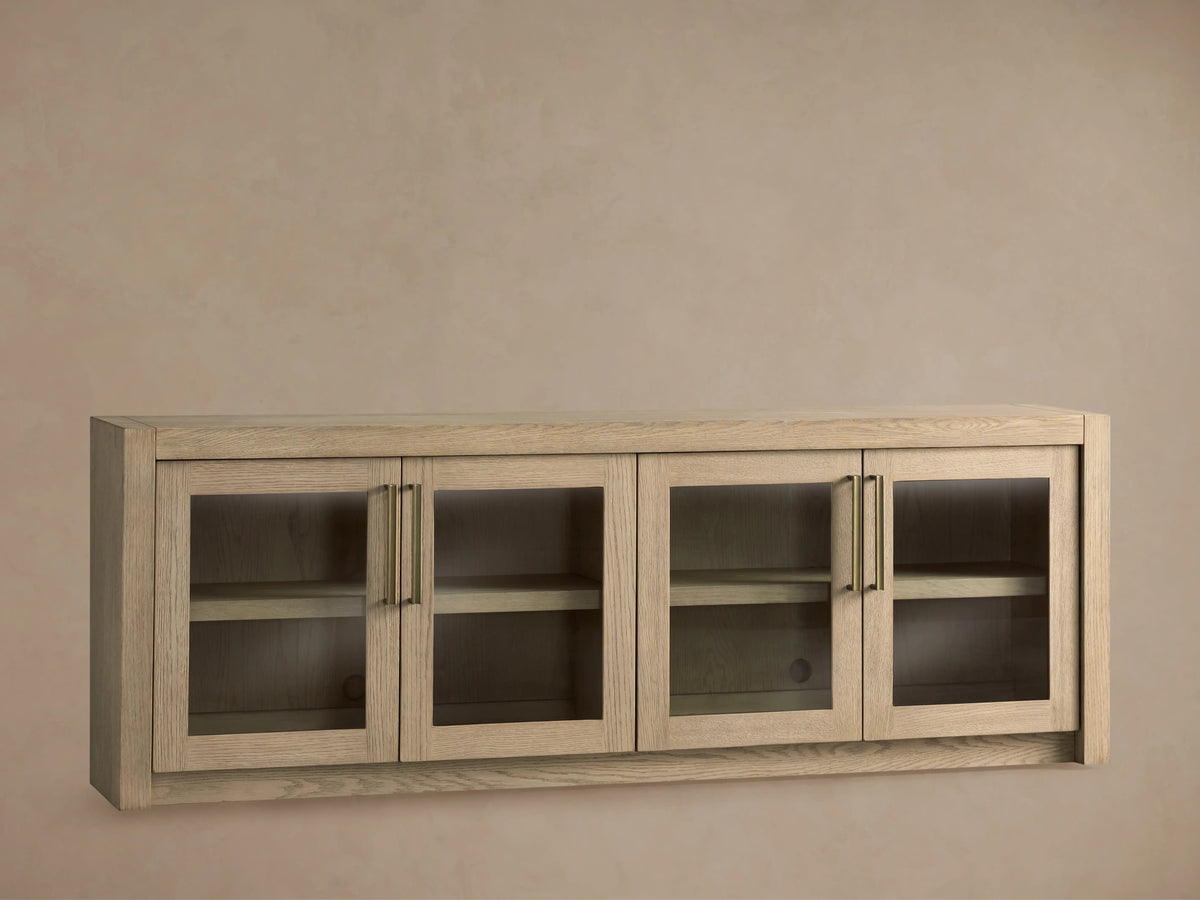 Bodhi Media Console in light oak, wall-mounted with four glass-panel doors, adjustable shelves, and brass handles on a beige background.
