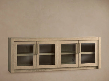 Bodhi Media Console in light oak, wall-mounted with four glass-panel doors, adjustable shelves, and brass handles on a beige background.