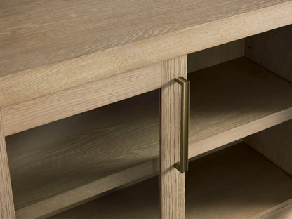 Bodhi Media Console with a solid white oak frame, four glass doors, brass handles, on a wood floor against a beige wall.
