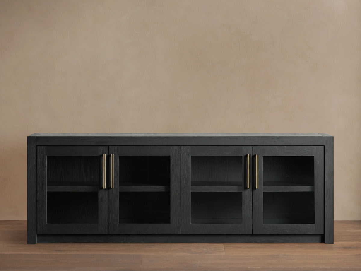 Bodhi Media Console in black oak with four glass-paneled doors, brass handles, on a wooden floor with a beige wall background.