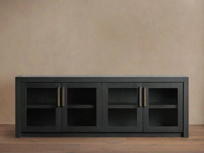 Bodhi Media Console in black oak with four glass-paneled doors, brass handles, on a wooden floor with a beige wall background.