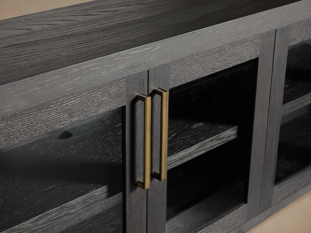 Bodhi Media Console in black oak with four glass-paneled doors, brass handles, on a wooden floor with a beige wall background.