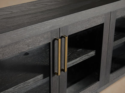 Bodhi Media Console in black oak with four glass-paneled doors, brass handles, on a wooden floor with a beige wall background.