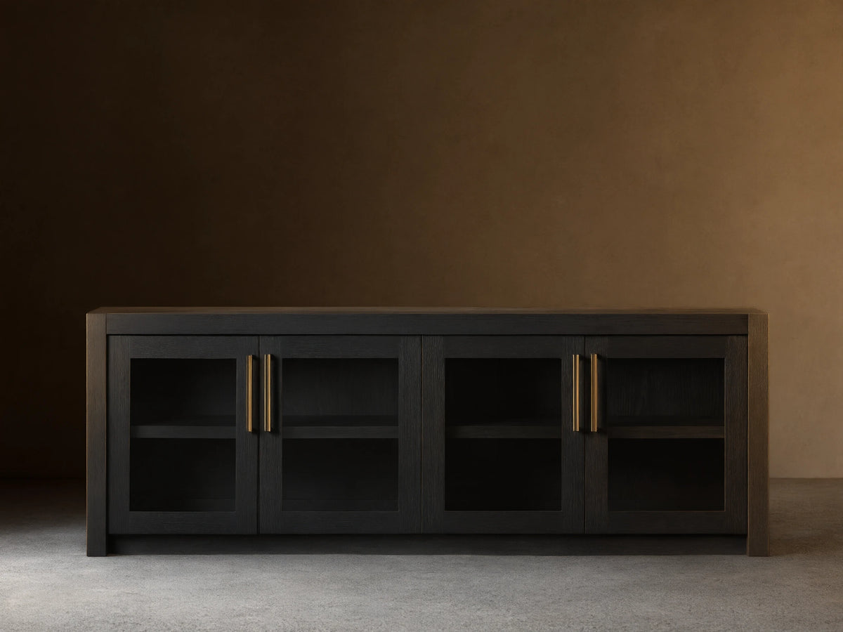 Bodhi Media Console in black oak with four glass-paneled doors, brass handles, on a wooden floor with a beige wall background.