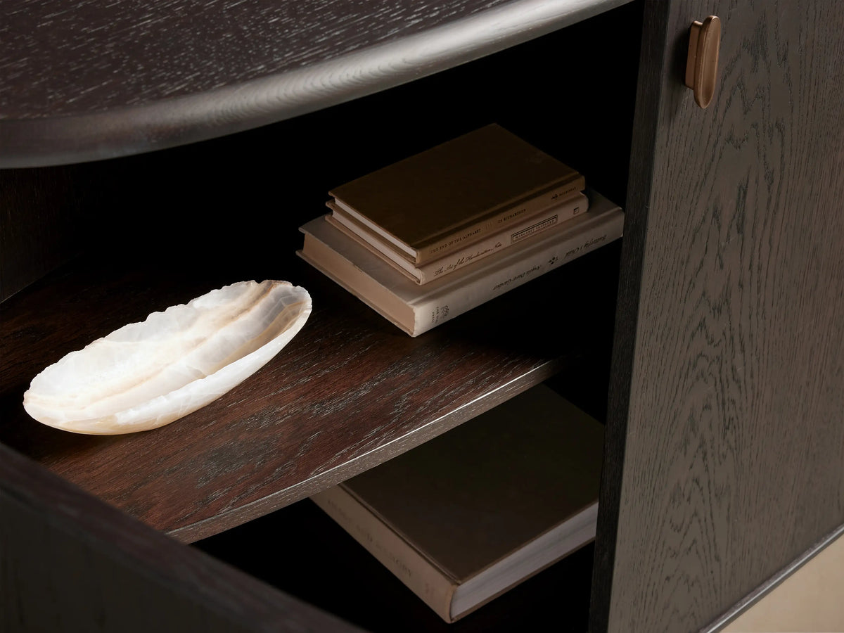 Dark Abanos Bexler Sideboard interior. Two wood shelves display books and a white onyx bowl. Door with antique bronze handle on right.
