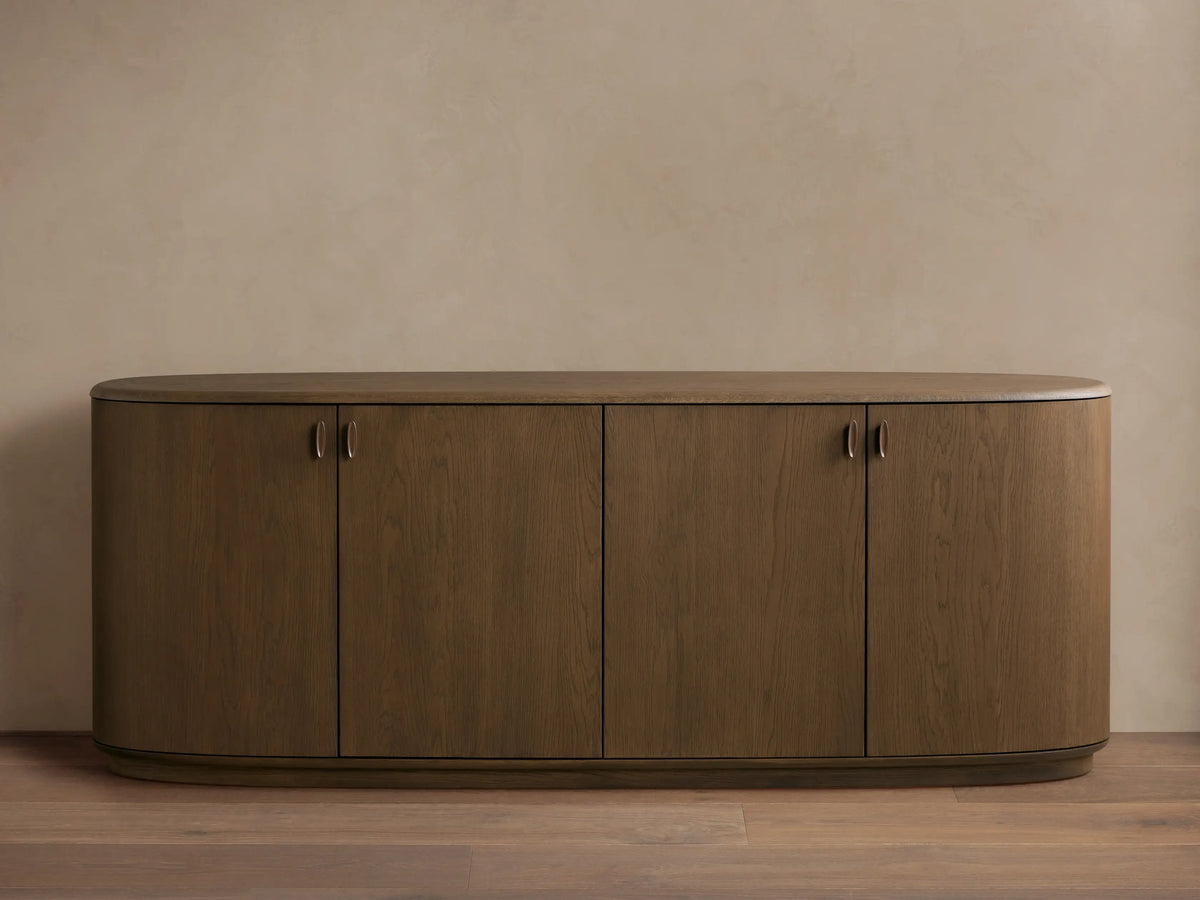 Bexler Sideboard, a walnut-finished wooden cabinet with rounded ends, an oval top, four doors, and small antique brass pulls.