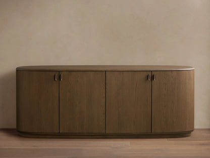 Bexler Sideboard, a walnut-finished wooden cabinet with rounded ends, an oval top, four doors, and small antique brass pulls.