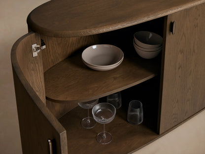 Close-up of open dark brown Bexler Sideboard, showing curved interior shelves with gray bowls, silver hinge, glasses & tumblers.