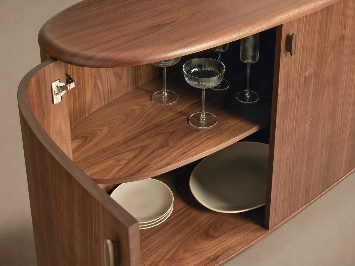 Close-up of a curved, dark walnut Bexler Sideboard with an open door revealing glass coupes, flutes, and light plates on interior shelves.
