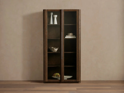 Dark wood Bardot Cabinet with two glass doors & bronze handles. Shelves hold decor items. Light wall & wood floor.